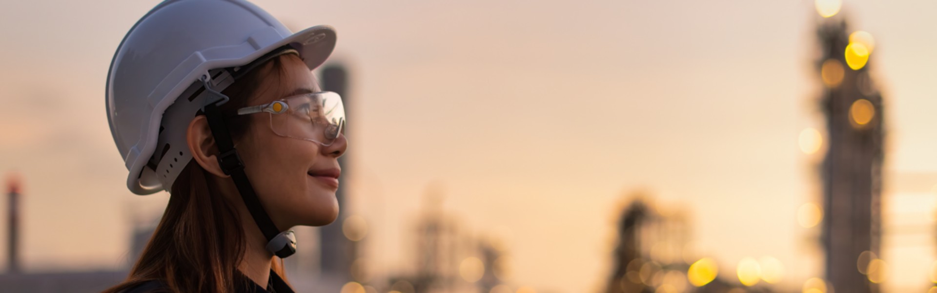 A woman in a safety helmet and goggles, looking confidently at an industrial site during sunset.