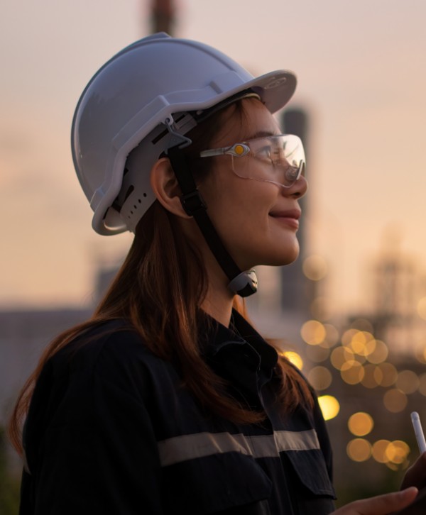 A woman in a safety helmet and goggles, looking confidently at an industrial site during sunset.