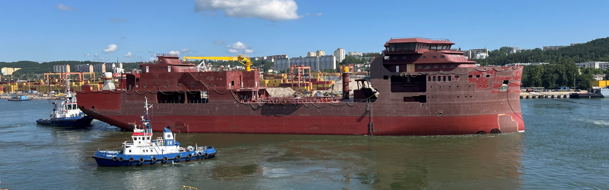 Large red ship being assisted by tugboats in a busy port.