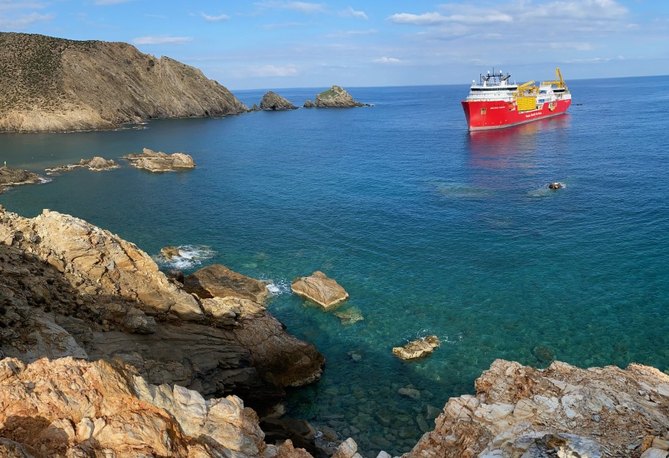 Coastal view with rocky cliffs and a large red ship in clear blue waters.