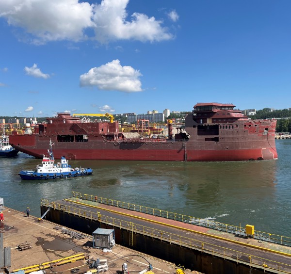 Grand navire rouge partiellement construit au port, avec un petit remorqueur à côté dans l'eau.