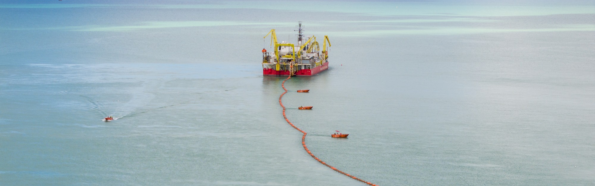 A large ship with yellow cranes and orange buoys in calm waters near the shore.
