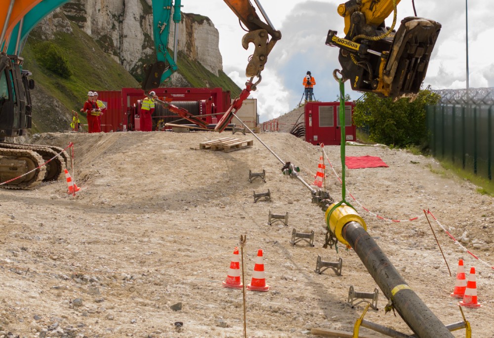 Construction site with machinery and workers laying a large pipe, surrounded by safety cones.