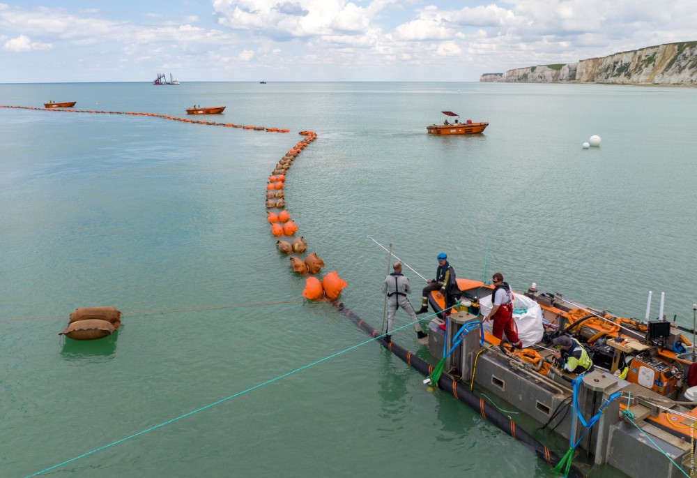 Crew members working on a floating barrier in calm waters with boats nearby and cliffs in the background.
