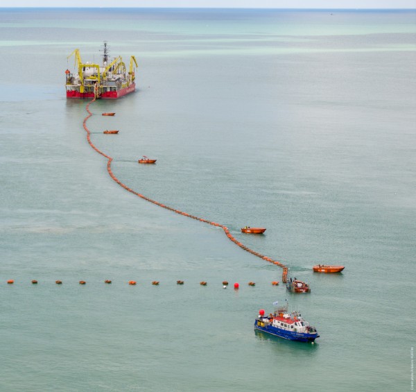 A large vessel with yellow cranes in the sea connected by a rope to smaller boats.