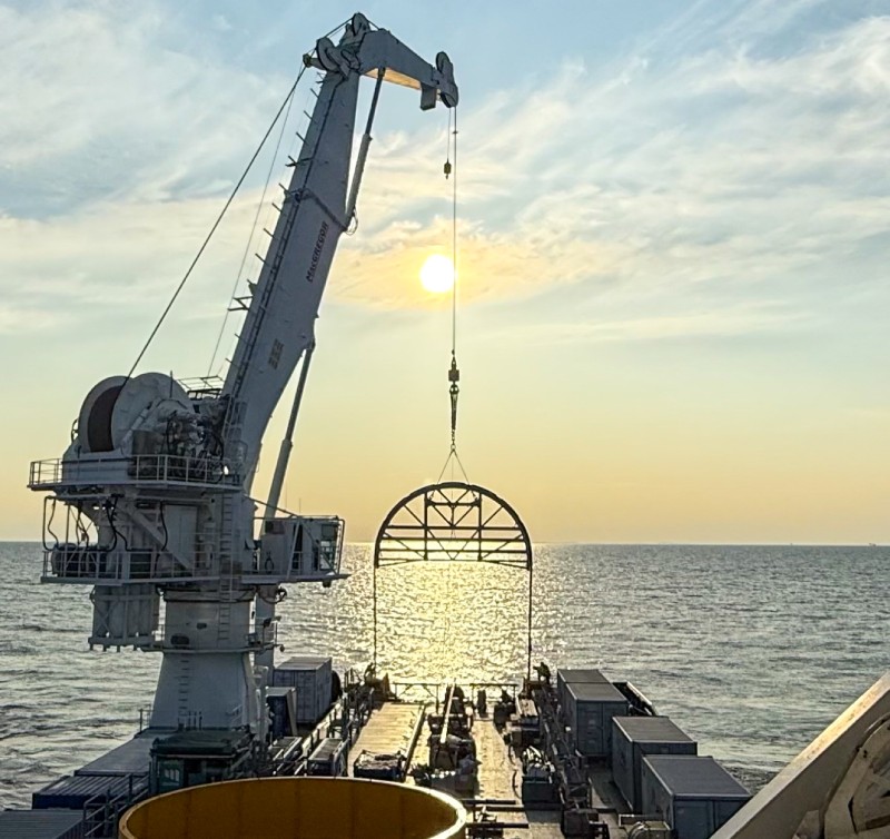 Crane lifting a circular structure over the ocean at sunset.